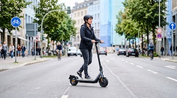 Una persona con casco y chaqueta oscura se desplaza en un scooter eléctrico sobre el asfalto de una calle con edificios al fondo.