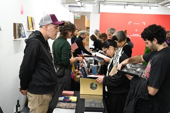 Grupo de personas hojeando libros y obras de arte en mesas de exhibición. Se ven paredes blancas y una pared roja con un gráfico en el fondo