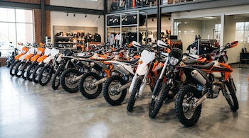 Fila de motocicletas deportivas de colores naranja, blanco y negro alineadas en el suelo de un concesionario con estantes de equipamiento al fondo.