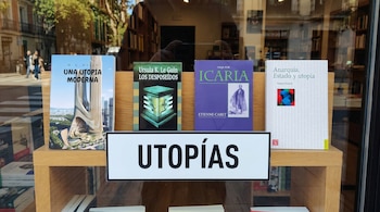 Cuatro libros y un cartel con "UTOPÍAS" sobre un estante de madera. Reflejos de una calle y estantes de librería en el cristal.