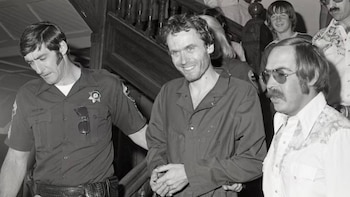 Fotografía en blanco y negro de Ted Bundy, con barba de unos días y pelo rizado, sonriendo mientras es escoltado por un oficial de policía y otro hombre