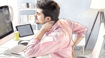 Un hombre de camisa rosa se toca el cuello y la zona lumbar. Está sentado en un escritorio con un monitor, auriculares, tablet, teclado y una lámpara.