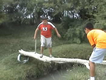 Un niño con camiseta roja camina sobre un tronco que cruza un arroyo, mientras otro niño con camiseta naranja sostiene el tronco