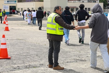 Hombre con chaleco amarillo guía a un grupo de personas en un área exterior pavimentada con conos naranjas hacia un edificio blanco con un letrero