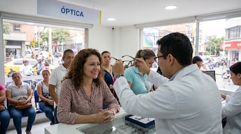 Un óptico en bata blanca muestra unas gafas negras a una mujer sonriente con cabello rizado en una óptica. Otras personas esperan en el fondo.