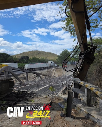 Brazo de excavadora con martillo hidráulico sobre escombros de concreto y varillas de metal de un puente, con camiones de carga al fondo