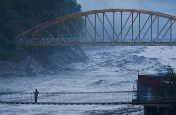 Un río de aguas grises y caudalosas fluye bajo un puente peatonal con una persona y un gran puente metálico amarillo