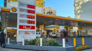 Una estación de servicio Shell con un cartel digital de precios en rojo, bombas de combustible amarillas y blancas, y dos furgonetas blancas estacionadas. Una persona camina a la izquierda