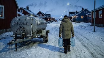 Una persona de espaldas con abrigo de invierno camina por la nieve con dos bidones de agua junto a un tanque cisterna y casas de madera.