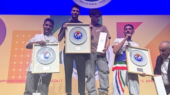 Cuatro hombres sonríen en un escenario con una pantalla azul y naranja de fondo. Sostienen premios: tres platos enmarcados y una caja blanca