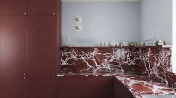 Cocina con gabinetes borgoña, encimera y salpicadero de mármol rojo veteado blanco, pared celeste, dos luces circulares, cristalería, platos y candelabros.