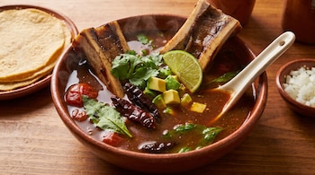 Un tazón de caldo humeante con dos huesos de médula, jitomates, chiles secos, cilantro, epazote, aguacate y lima. Al lado, tortillas y cebolla picada.