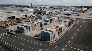 Filas de contenedores de carga, camiones de servicio y aviones estacionados frente a depósitos en la pista de un aeropuerto.