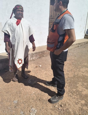 Un hombre con vestimenta tradicional indígena (poncho blanco, pañuelo rojo, pies descalzos) y un hombre con chaleco salvavidas naranja están de pie en tierra