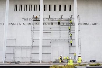 Fachada de edificio de mármol blanco con ventanas y letras. Andamios de metal cubren parte de la pared. Varios trabajadores con chalecos amarillos y cascos