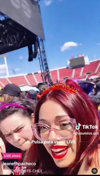 Una joven de cabello rojo con anteojos rosa y otra persona están frente a la cámara en un estadio con gradas rojas y un escenario al fondo