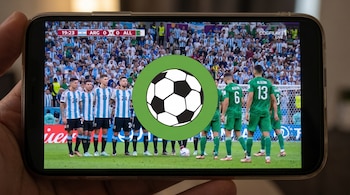 Primer plano de una mano sosteniendo un smartphone que muestra un partido de fútbol entre Argentina (azul y blanco) y Argelia (verde) con un logo de balón en el centro.