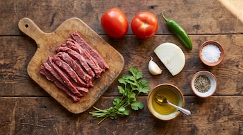 Tiras de carne cruda en una tabla, dos tomates, un chile verde, cebolla, ajo, cilantro, aceite, sal y pimienta sobre una mesa de madera.