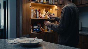 Una persona busca comida dentro de un armario de cocina abierto y con estantes llenos de paquetes, frente a un plato en la encimera