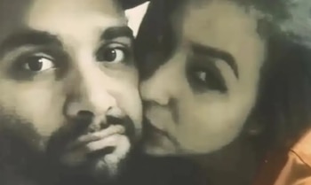 Primer plano de una pareja. Un hombre con barba mira a la cámara, mientras una mujer de cabello oscuro, parcialmente visible, apoya su mejilla en la suya