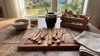 Mesa de madera con bastones de pan con semillas sobre una tabla, un mate con bombilla, un tazón con semillas y una pequeña caja.