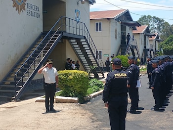 Oficiales de la Policía Nacional Civil en uniformes claro y oscuro saludan durante una formación exterior frente a edificios de estación con escaleras metálicas