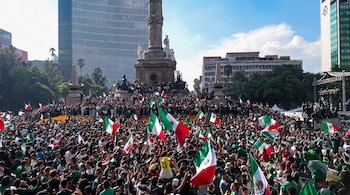 Multitud de personas que portan banderas de México. Al fondo, el monumento del Ángel de la Independencia, edificios y cielo azul con nubes.