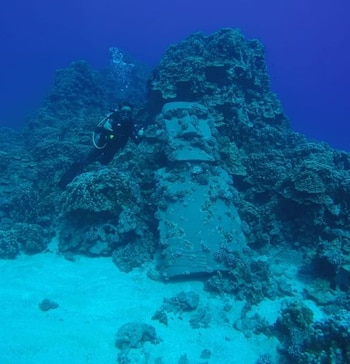 Buceador con equipo de inmersión, estatua Moai sumergida, arrecife de coral, fondo marino arenoso, agua azul