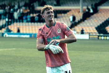 Bobby Faulkner sonriendo en la cancha de fútbol con camiseta rosa estampada, pantalón corto blanco y guantes de arquero