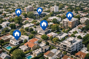 Vista aérea de un barrio residencial con casas, edificios, árboles y calles. Varios marcadores azules con íconos de casas blancas resaltan propiedades.