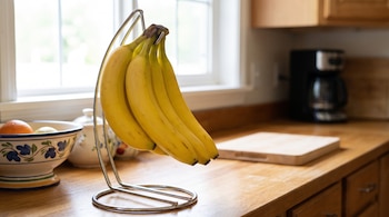 Racimo de bananas amarillas colgado en un soporte de metal sobre una mesada de madera junto a un frutero y una cafetera.