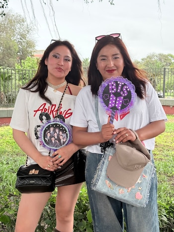 Dos jóvenes sonríen al aire libre mientras sostienen abanicos artesanales con imágenes y el logotipo del grupo de música BTS