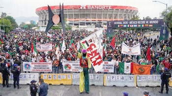 Multitud protesta en calle frente al Estadio Banorte. Pancartas sobre desaparecidos y banderas de México. Persona en zancos, atuendo vibrante. Banner del Mundial 2026 y policía.