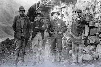 Cuatro hombres posan al aire libre, tres con sombreros y uno con gorra y poncho, frente a muros de piedra. La foto es en blanco y negro y muestra ropa de principios del siglo XX