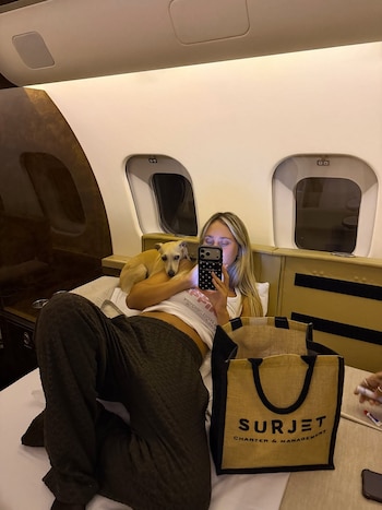Mujer rubia recostada en un jet privado con un perro galgo italiano, mirando un teléfono móvil. A su lado, una bolsa con el logo Surjet