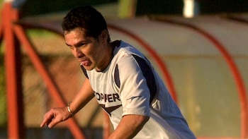 Primer plano de Hugo Morales, hombre, sudoroso y con la lengua ligeramente fuera, vistiendo camiseta deportiva clara con detalles oscuros. Apunta con el dedo índice derecho. Fondo borroso