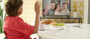 Una mujer mayor en una camisa roja levanta su mano hacia una pantalla de televisión que muestra a una pareja joven con un bebé saludando