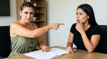 Dos mujeres en oficina. Una, de verde, frunce el ceño y señala a la otra, de negro, que luce asombrada y preocupada.