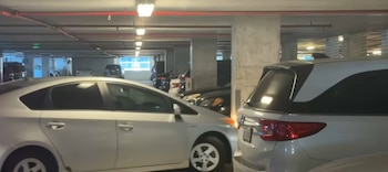 Imagen de un estacionamiento subterráneo con varios coches, incluyendo un sedán plateado y un SUV blanco, bajo luces fluorescentes y tuberías rojas