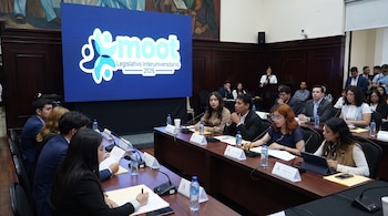 Jóvenes sentados en mesas con micrófonos y botellas de agua en un salón formal. Pantalla grande con el logo 'Moot Legislativo Interuniversitario 2026' al fondo.