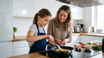 Una niña con delantal cocina en una sartén junto a una mujer, sobre una encimera de madera con cáscaras de huevo y verduras picadas.