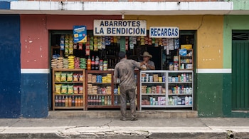 Fachada colorida de una tienda en El Salvador con letrero "Abarrotes La Bendición", mostrando estantes con snacks, bebidas y artículos; dos personas no identificables están dentro.