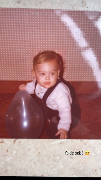 Un bebé de cabello claro, vestido con un suéter blanco y peto oscuro, sentado junto a un globo gris. El fondo es una pared rosa con lunares blancos