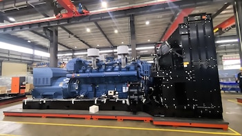 Un generador eléctrico industrial de gran tamaño, de color azul y negro, se muestra en el suelo de una fábrica con techo alto y grúas