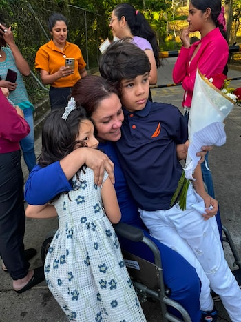 Mujer en silla de ruedas abraza a una niña con tiara y un niño que sostiene un ramo de flores. Varias personas observan alrededor