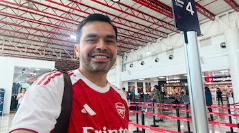 Primer plano de un hombre sonriendo con camiseta roja y blanca del Arsenal en un aeropuerto. Se ven personas, asientos, tiendas y un cartel de Zona 4 al fondo.