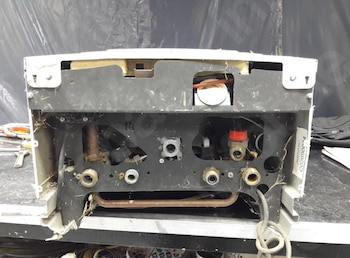 Parte inferior desarmada de un calentador con tuberías de cobre, conectores metálicos y cables visibles sobre un banco de trabajo