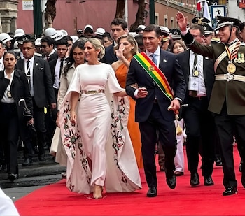 Un hombre con traje y banda tricolor camina junto a una mujer con vestido blanco sobre una alfombra roja, acompañados por guardias