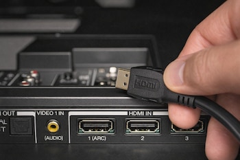 Primer plano de una mano sosteniendo un cable HDMI negro y dorado a punto de conectarlo al puerto HDMI 1 (ARC) en la parte trasera de un dispositivo electrónico.