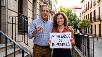 Un hombre y una mujer sostienen un juego de llaves y un cartel con el texto PROPIETARIOS DE INMUEBLES en una calle.